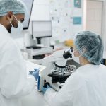 Clinicians in white coats; one is standing next to the other who is looking int a microscope.