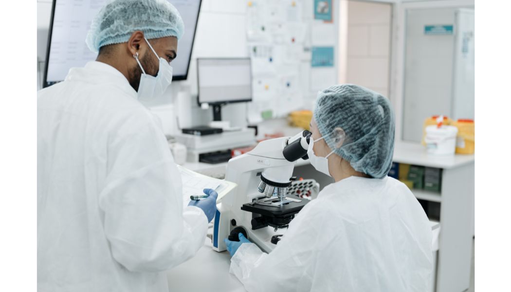 Clinicians in white coats; one is standing next to the other who is looking int a microscope.
