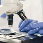 Hands in blue gloves setting a slide under a microscope